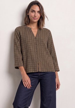 Femme portant une chemise marron à carreaux boutonnée avec manches trois-quarts et un jean bleu foncé sur un fond clair uni.