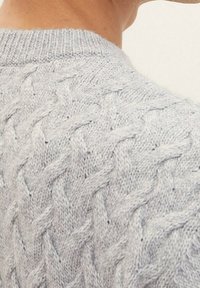 Pull gris clair en tricot avec un motif torsadé. L'encolure est côtelée, avec une vue en gros plan du pointage complexe.