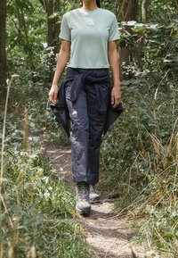 Camiseta corta de color verde claro, pantalones negros cónicos para exteriores atados en la cintura y botas de senderismo grises, usados en un sendero forestal.