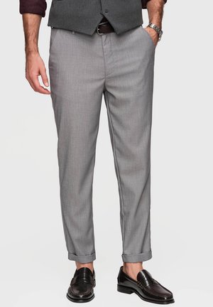 Homme portant un pantalon de costume gris clair avec des ourlets retroussés, des mocassins en cuir noir, un gilet gris foncé et une chemise bordeaux avec les manches remontées.