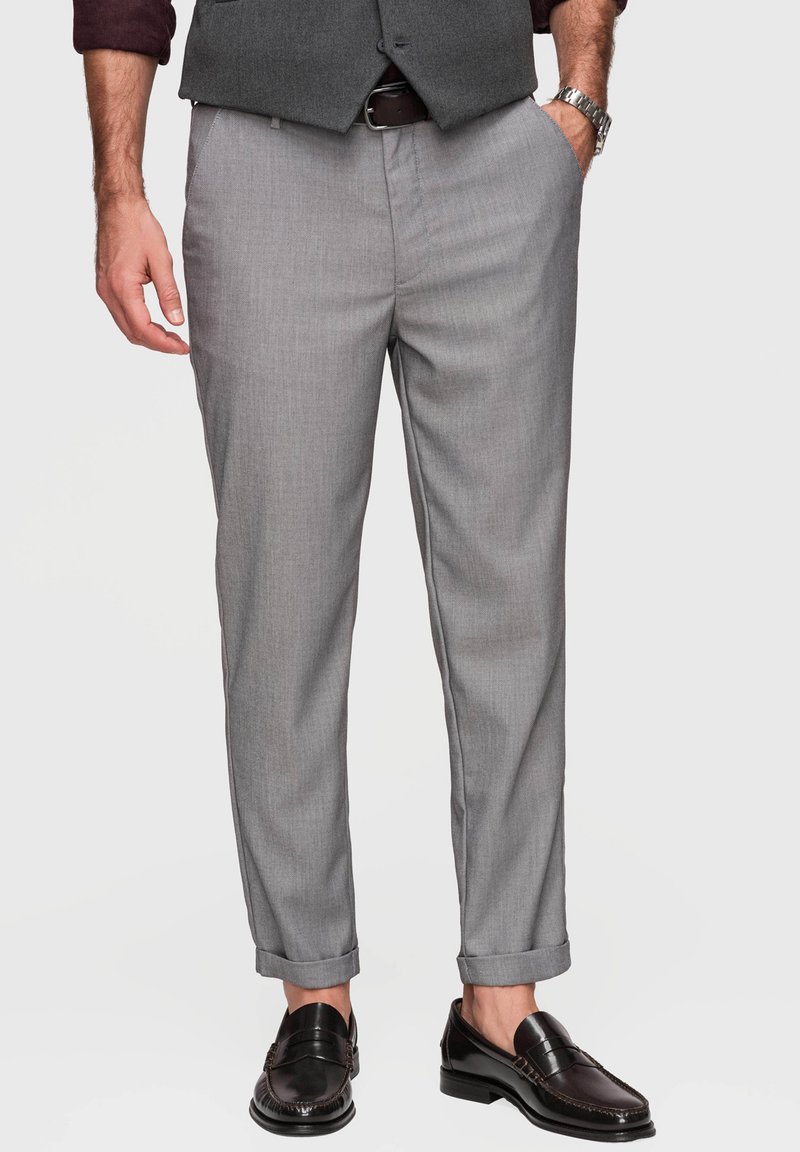 Homme portant un pantalon de costume gris clair avec des ourlets retroussés, des mocassins en cuir noir, un gilet gris foncé et une chemise bordeaux avec les manches remontées.