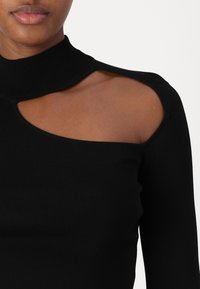 Haut noir à manches longues côtelé avec un col montant et un design découpé sur l'épaule gauche, mettant en valeur un tissu texturé et une forme ajustée.