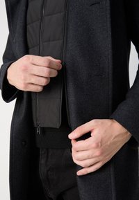 Manteau en laine sombre avec une finition lisse et texturée. Il présente une couche intérieure matelassée zippée en noir, montrée avec des mains ajustant la fermeture éclair.