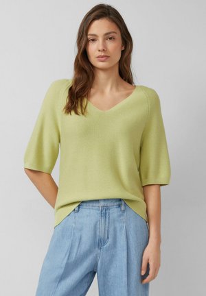 Frau mit langem braunem Haar, die einen hellgrünen V-Ausschnitt-Pullover mit Ellbogen-langen Ärmeln und hellblaue, hoch taillierte Jeans trägt.