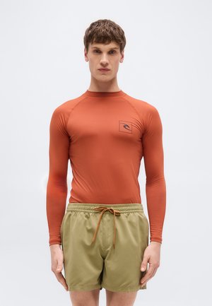 Mężczyzna ubrany w długi rękaw koszulki termicznej w kolorze rdzy oraz oliwkowe zielone szorty do pływania z troczkiem na tle jednolitym.