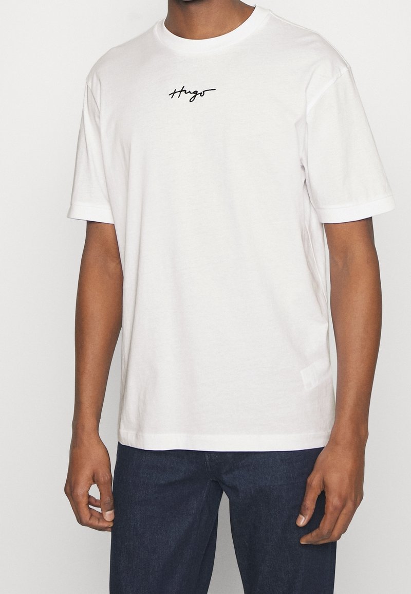 T-shirt en coton blanc à manches courtes, avec un logo "Hugo" en cursive noire sur la poitrine. Encolure ronde simple et coupe décontractée.
