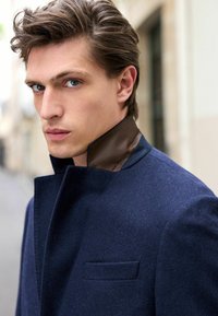 Manteau en laine bleu marine avec un col haut, présentant une accentuation texturée marron. Le manteau comprend une poche poitrine unique et une silhouette ajustée.