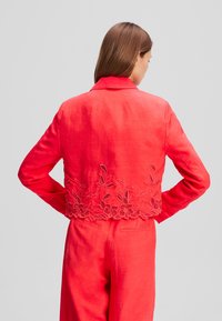 Rote Leinen-Cropped-Jacke mit Kragen, verziert mit floraler Stickerei am Saum, langen Ärmeln und einer glatten Textur.