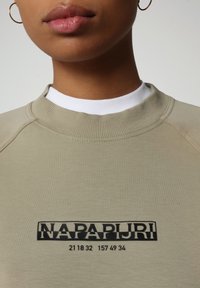 Sudadera color beige claro con una tela texturizada, que presenta un logo y números impresos en negro. Cuello redondo y puños acanalados.