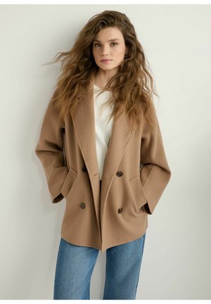 Kobieta z długimi falującymi włosami, ubrana w beżowy dwurzędowy płaszcz na białą bluzkę i niebieskie dżinsy, stojąca na jednolitym jasnym tle.