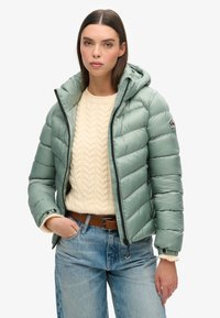 Chaqueta acolchada verde menta con capucha, que presenta costuras horizontales, cierre de cremallera y dos bolsillos laterales, llevada sobre un suéter de punto trenzado color crema.