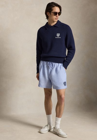 Άνδρας που φοράει μπλε πουλόβερ Wimbledon, ανοιχτά μπλε σορτς, λευκές κάλτσες, αθλητικά παπούτσια και γυαλιά ηλίου, στέκεται μπροστά σε ένα απλό μπεζ φόντο.