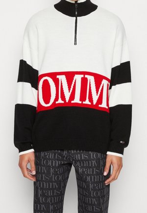 Person trägt einen schwarz-weiß-roten Strickpullover mit halbem Reißverschluss und auffälligem "TOMMY" Schriftzug, kombiniert mit dunklen Jeans mit wiederholtem "tommy jeans" Druck.