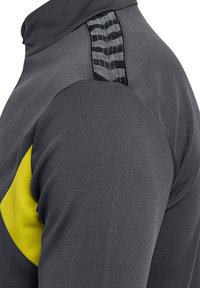 Graues Sportoberteil mit gelben Akzenten, das über eine strukturierte Oberfläche, Netzpaneele und ein einzigartiges schwarzes Musterdetail an der Schulter verfügt.