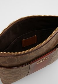 Brązowa torba z wzorzystego materiału z złotym suwakiem; wnętrze ma brązowe wykończenie oraz naszywkę z informacjami o marce. Posiada skórzany akcent.