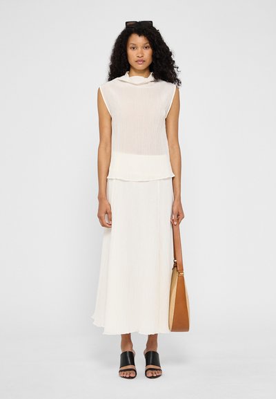 Débardeur blanc plissé sans manches superposé sur une jupe longue assortie. Sandales noires à glissière et un sac à épaule marron à texture lisse.