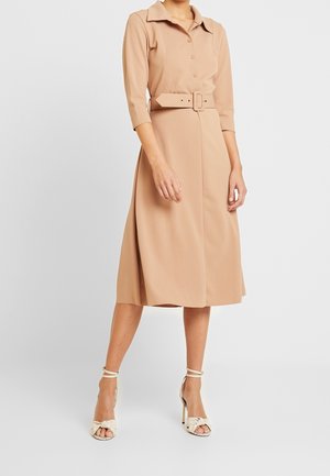 Femme portant une robe beige ceinturée, longueur genoux, avec des manches trois-quarts et des sandales blanches à talons hauts avec brides à la cheville.