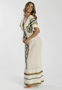 Vestido maxi de tela crema con patrones en forma de zigzag en negro, dorado y verde. Presenta un escote en V, mangas cortas y un dobladillo con volantes.