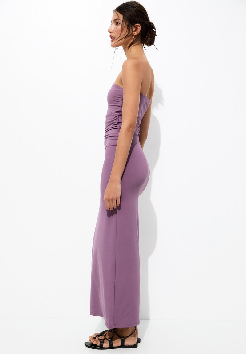 Robe maxi sans bretelles violette avec des détails froncés au niveau du corsage, texture lisse et coupe ajustée, associée à des sandales noires à lanières.