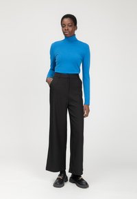 Maglione a collo alto blu aderente abbinato a pantaloni neri a vita alta e gamba larga, con scarpe nere chunky con accenti dorati.