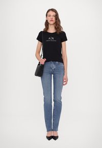 Tricou din bumbac negru cu logo-ul "A|X ARMANI EXCHANGE", asortat cu blugi direcți de culoare albastră și pantofi cu toc înalt negri. Modelul ține de asemenea o geantă mică neagră.