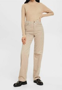 Woman wearing a beige ribbed long-sleeve top and high-waisted beige corduroy trousers paired with black loafers, standing with one hand on hip.