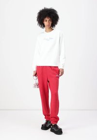 Hanorac alb cu logo albastru, pantaloni roșii de trening și pantofi cu talpă înaltă negri. Modelul ține o geantă mică roz. Texturi netede, design casual.