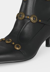 Botte à talon en cuir noir avec bout pointu ornée de clous ronds embossés dorés et noirs sur des sangles à l'avant.