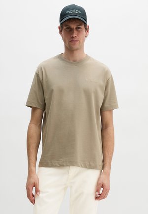 Mann trägt ein beigefarbenes Kurzarm-T-Shirt von Marc O’Polo, off-white Hose und eine dunkelblaue Marc O’Polo-Kappe, steht vor einem weißen Hintergrund.