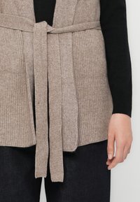 Beige gebreide cardigan met een ribbelstructuur, open voorkant en een bijpassende strikriem. Draagt over een zwarte top en donkere jeans.