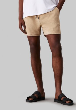 Hombre con pantalones cortos beige de tela tipo toalla con cordón, camiseta blanca y sandalias negras sin cordones sobre fondo gris.