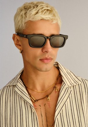 Homme aux cheveux courts décolorés portant des lunettes de soleil noires, un anneau d'oreille en or, des colliers superposés et une chemise blanche à rayures verticales noires.
