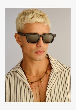 Mann mit kurz gebleichtem Haar, der schwarze Sonnenbrille, goldenen Creolen, mehrere Halsketten und ein weißes Hemd mit schwarzen vertikalen Streifen trägt.