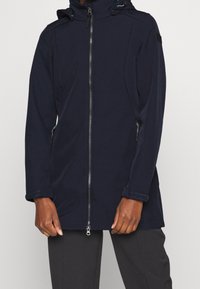 Giacca softshell blu navy con zip frontale, cappuccio regolabile e tasche laterali. Presenta un design elegante, polsini testurizzati e accessori minimali.