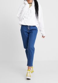 Bílá mikina s kapucí a šňůrkami, kombinovaná s modrými džínami. Žluté tenisky doplňují outfit kontrastními tkaničkami a klasick�ým designem.