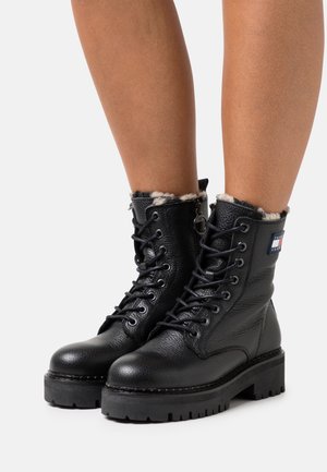 Botas de cuero negro hasta el tobillo con cordones, forro interior acolchado, suela negra resistente y detalle de logo en el lateral. Acabado texturizado, altura hasta la pantorrilla.