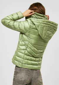 En person justerer hætten på en lysegrøn, quiltet jakke, iført grå slidte jeans, vist fra ryggen mod en ensfarvet baggrund.