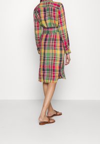 Frau in einem bunten karierten Kleid mit Gürtel und braunen flachen Sandalen, stehend vor einem schlichten weißen Hintergrund, mit dem Rücken zur Kamera.