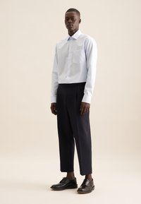 Camicetta button-up a righe azzurre chiaro con taschino sul petto, abbinata a pantaloni larghi scuri e scarpe nere stringate. Tessuto liscio e vestibilità sartoriale.