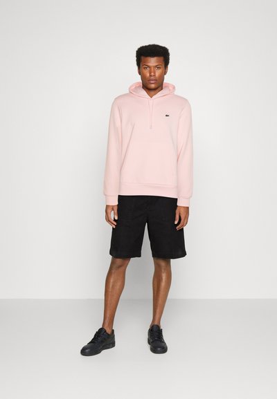 Sweatshirt à capuche rose clair avec une poche kangourou, associé à un short noir. Des chaussures noires complètent la tenue. Design simple et décontracté.