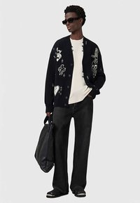 Muž v černém cardigan s bílými grafickými vzory, bílou košili, černých širokých kalhotách, slunečních brýlích a s velkou černou taškou.