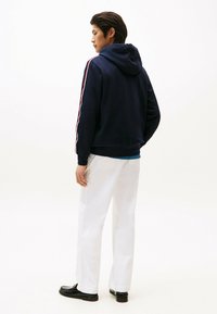 Granatowy hoodie ze sznurkiem, białe i czerwone paski na ramionach, w połączeniu z białymi spodniami i czarnymi loafersami, pokazany od tyłu.