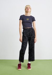 Pantaloni neri a gamba larga con una texture a fantasia, abbinati a una maglietta blu marina aderente con una grafica colorata. Scarpe sportive bordeaux, calze sovrapposte.