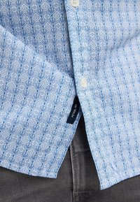 Camicia azzurra a fantasia con design geometrici, dotata di abbottonatura frontale, bottoni bianchi e un'etichetta del marchio visibile sulla cucitura laterale.