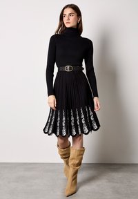 Zwarte geribbelde jurk met een col, uitgerust met een uitlopende rok met witte bloemenborduursels aan de zoom en een zwarte riem die de taille accentueert.