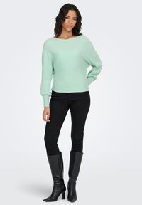 Maglione verde menta a coste con scollo ampio e maniche lunghe, abbinato a jeans skinny neri e stivali neri al ginocchio.