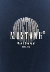 Sötétkék pamut póló, amelyen egy nagy, textúrált ezüst logó található, alatta fehér betűkkel: "MUSTANG JEANS COMPANY" és "SINCE 1932".