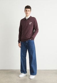 Bordo zip-up dukserica s logotipom štita, uparena s labavim plavim trapericama i bijelim tenisicama, stoji na neutralnoj pozadini.