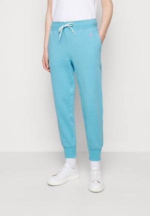 Femme portant un pantalon de jogging bleu clair avec cordon de serrage et logo rose, baskets blanches et chaussettes blanches, debout devant un fond uni.