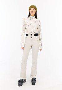 Svetlá dlhá blúzka s farebnými vzormi, spárovaná s krémovými tesnými nohavicami s čiernymi akcentmi a zipsovými vreckami; čierne lyžiarske topánky.